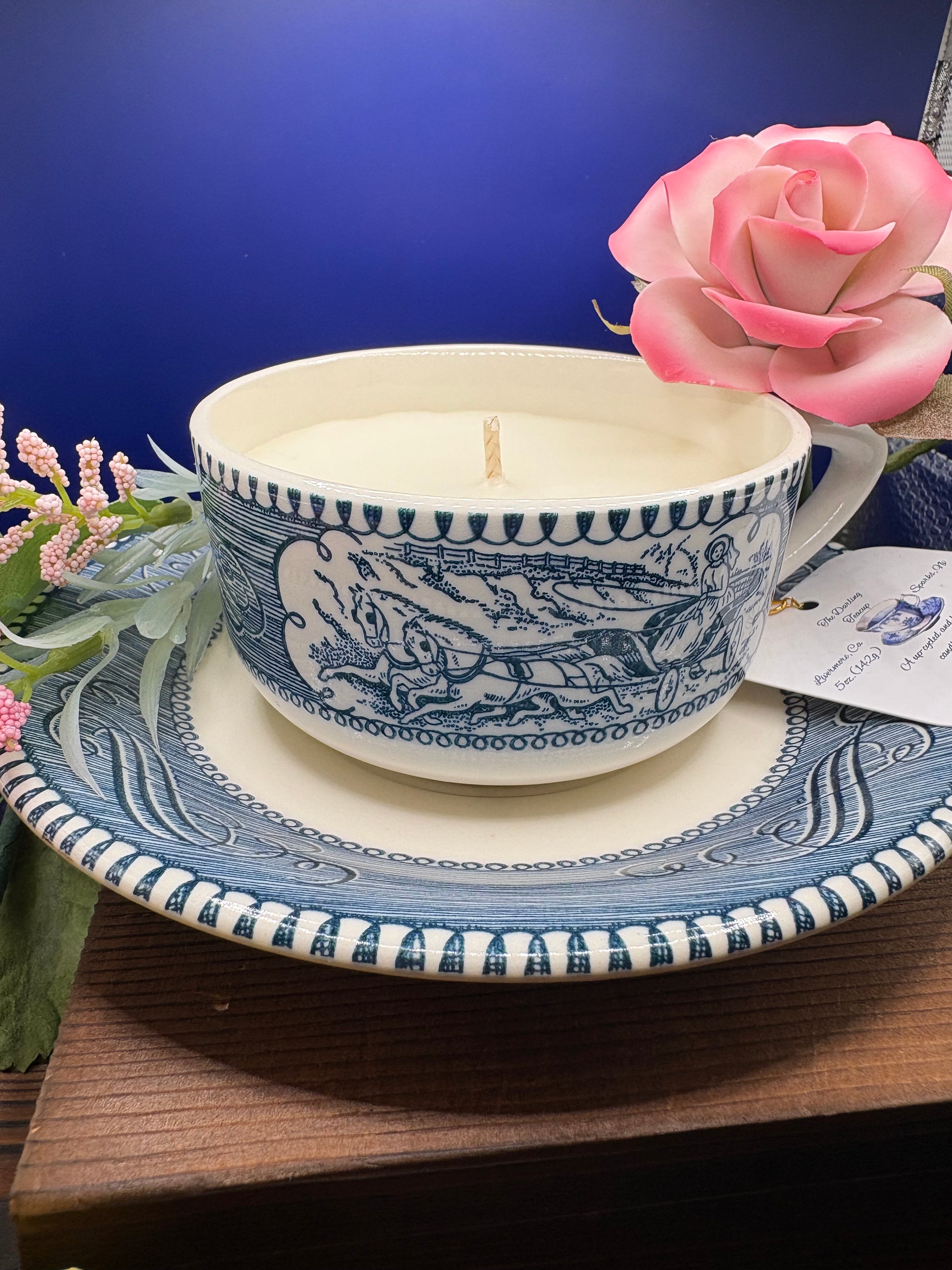 A blue and white Currier and Ives Teacup candle, displayed on a matching saucer and plate. The candle features a detailed blue landscape illustration on a cream-colored body.