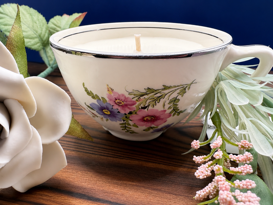 A  candle in a teacup-shaped cup with a floral print of pink, purple, and green flowers, set on a wooden surface with decorative white and pink flowers.