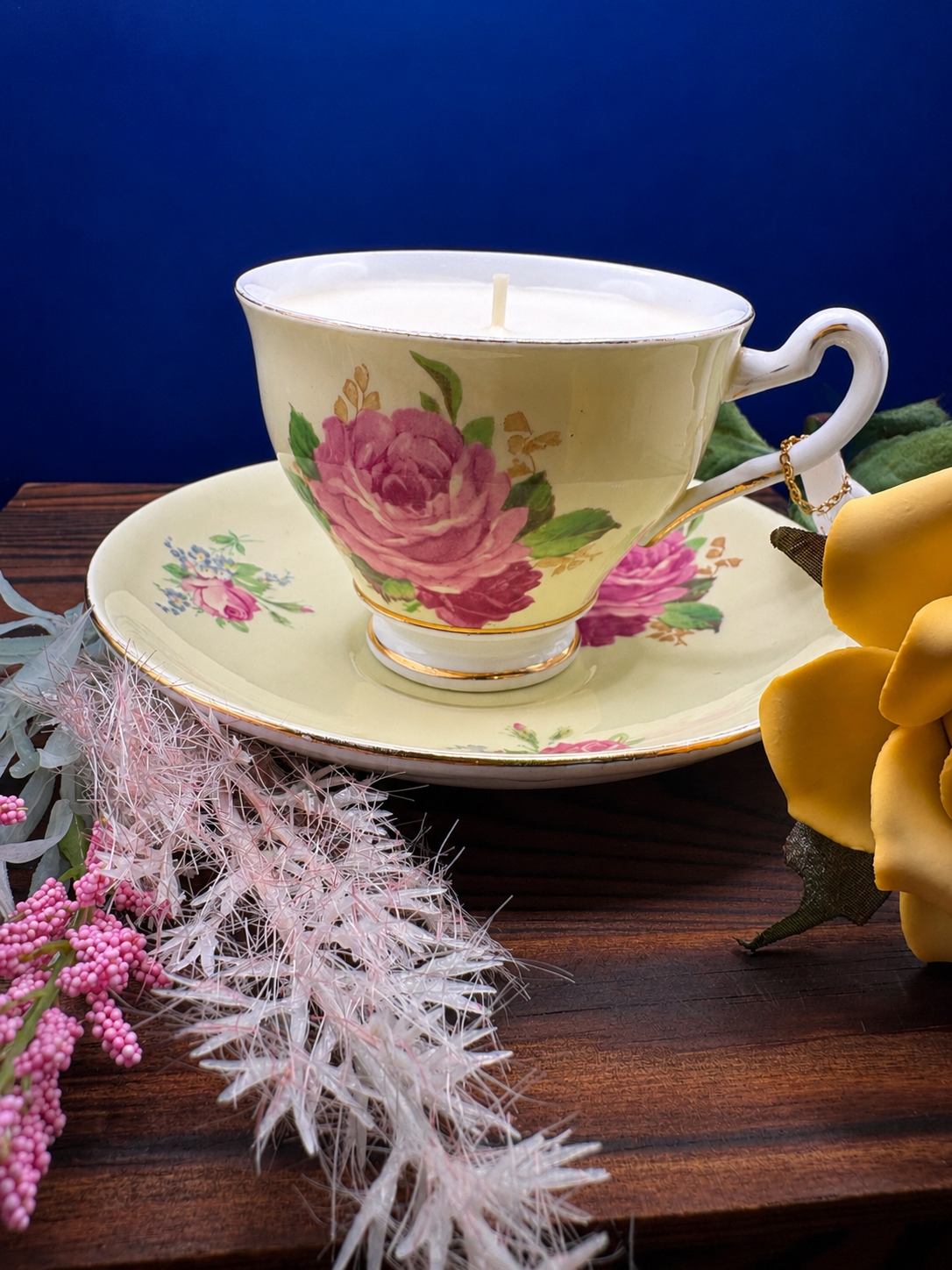 A floral teacup candle in a yellow teacup with pink roses and green leaves, resting on a matching saucer.
